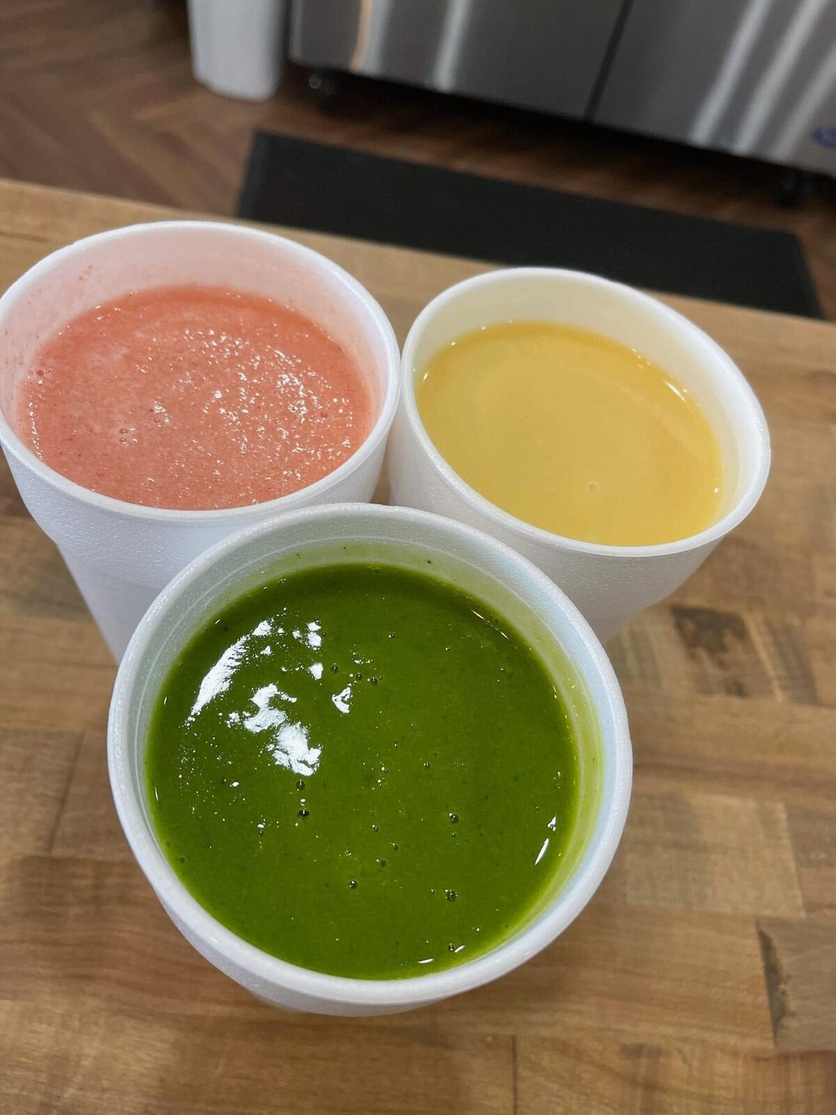 Three smoothies in white cups on the wood counter — pink, green and yellow